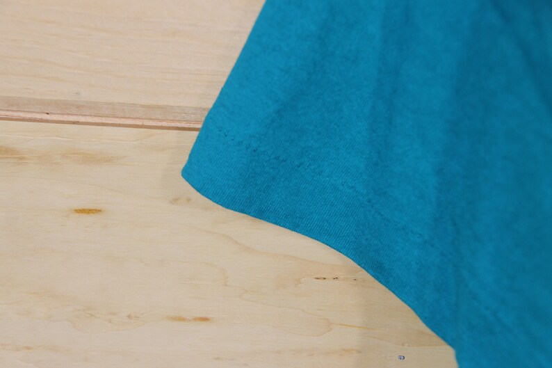 May include: Close-up of the turquoise blue fabric of a t-shirt. The fabric is smooth and has a slight sheen.