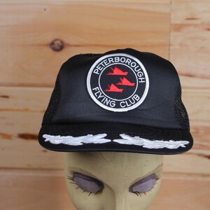 May include: Black trucker cap with a white embroidered design along the brim. The cap features a circular patch with the text "PETERBOROUGH FLYING CLUB" around a red graphic of three aeroplanes.