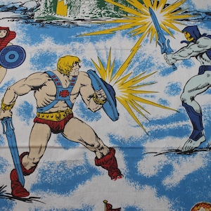 May include: A blue fabric featuring a printed illustration of He-Man, Skeletor, and Evil-Lyn in a battle scene. The characters are depicted with swords and shields against a sky-blue background with clouds, yellow starbursts, and planets.