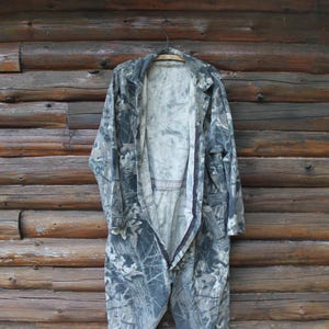 May include: A camouflage jumpsuit hangs on a wooden hanger against a rustic wooden wall. The jumpsuit is open, revealing the interior lining. The fabric features a mix of greens, greys, and browns, creating a woodland camouflage design.