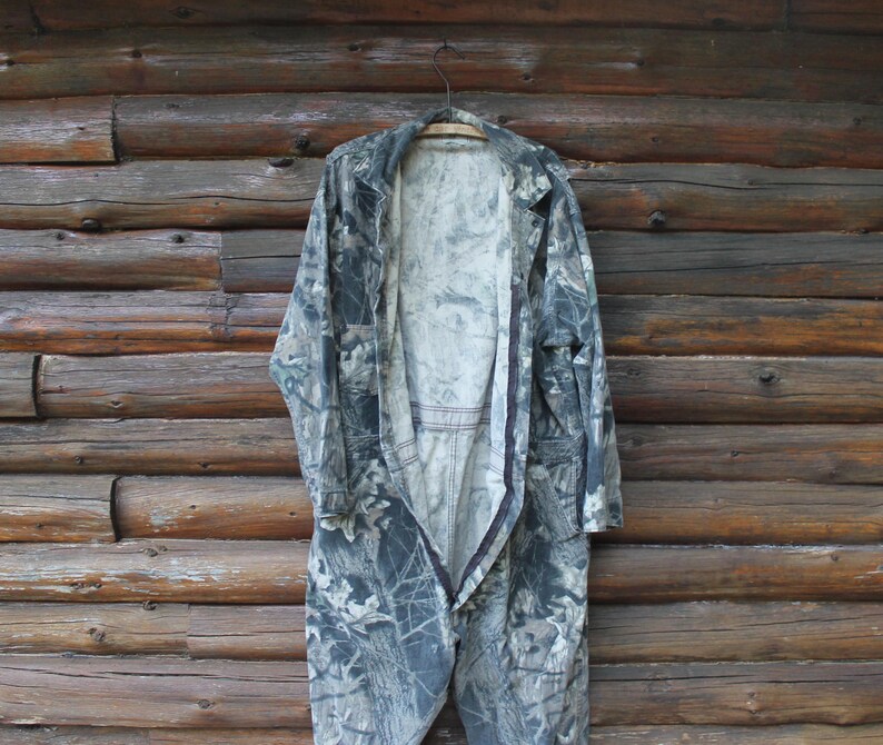 May include: A camouflage jumpsuit hangs on a wooden hanger against a rustic wooden wall. The jumpsuit is open, revealing the interior lining. The fabric features a mix of greens, greys, and browns, creating a woodland camouflage design.
