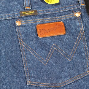 May include: A pair of blue denim jeans with a brown leather Wrangler label on the back pocket. The pocket has a distinctive W-shaped stitching.