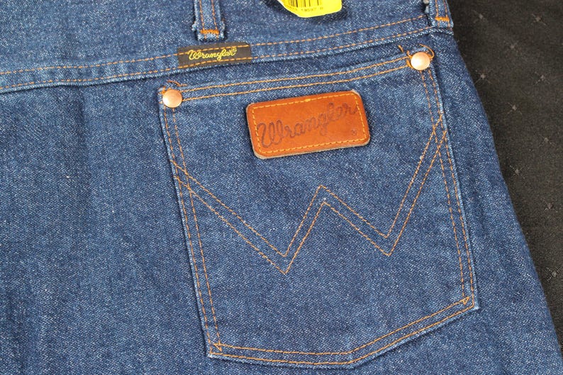 May include: A pair of blue denim jeans with a brown leather Wrangler label on the back pocket. The pocket has a distinctive W-shaped stitching.