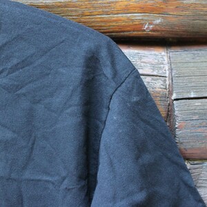 May include: A solid black t-shirt featuring a ribbed crew neckline and short sleeves. The fabric has a visible texture with natural folds and creases across the shoulder and chest area, showcasing the plain dark cotton material construction.