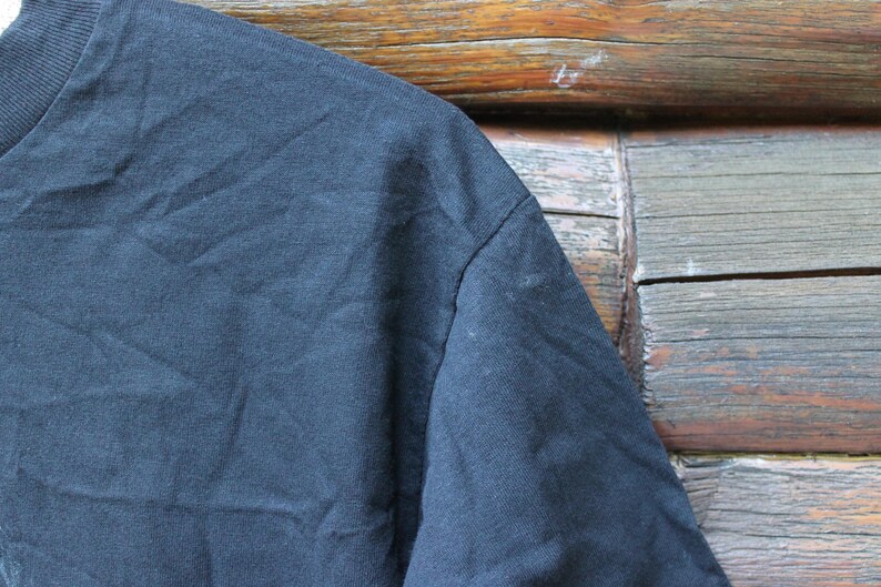 May include: A solid black t-shirt featuring a ribbed crew neckline and short sleeves. The fabric has a visible texture with natural folds and creases across the shoulder and chest area, showcasing the plain dark cotton material construction.