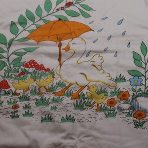 May include: A white fabric with a colorful illustration of a duck family walking under an orange umbrella in a field of green grass, mushrooms, and flowers. The duck family is yellow and white, and the flowers are red, blue, and yellow.