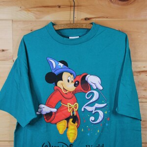 May include: Teal blue t-shirt featuring Mickey Mouse dressed as a sorcerer with a blue hat and red robe. Mickey is holding a wand and pointing it at the number 25. The text "Walt Disney World" is at the bottom of the shirt.