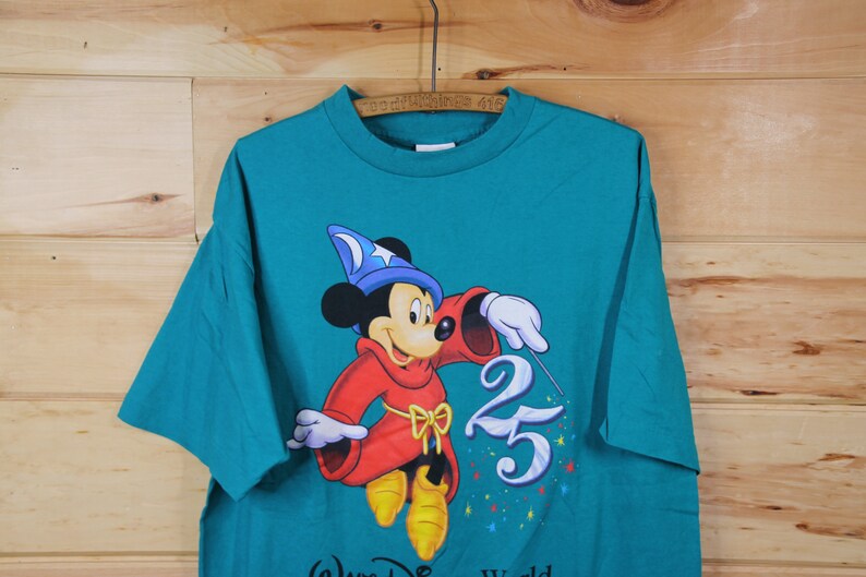 May include: Teal blue t-shirt featuring Mickey Mouse dressed as a sorcerer with a blue hat and red robe. Mickey is holding a wand and pointing it at the number 25. The text "Walt Disney World" is at the bottom of the shirt.