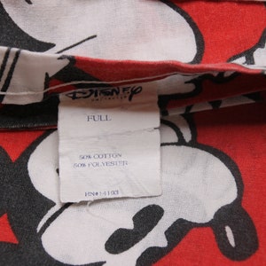 Op de afbeelding: Een wit label met zwarte tekst die "Disney", "Full", "50% Katoen 50% Polyester" en "RN#14103" luidt, tegen een rode en witte achtergrond met een Mickey Mouse-print.