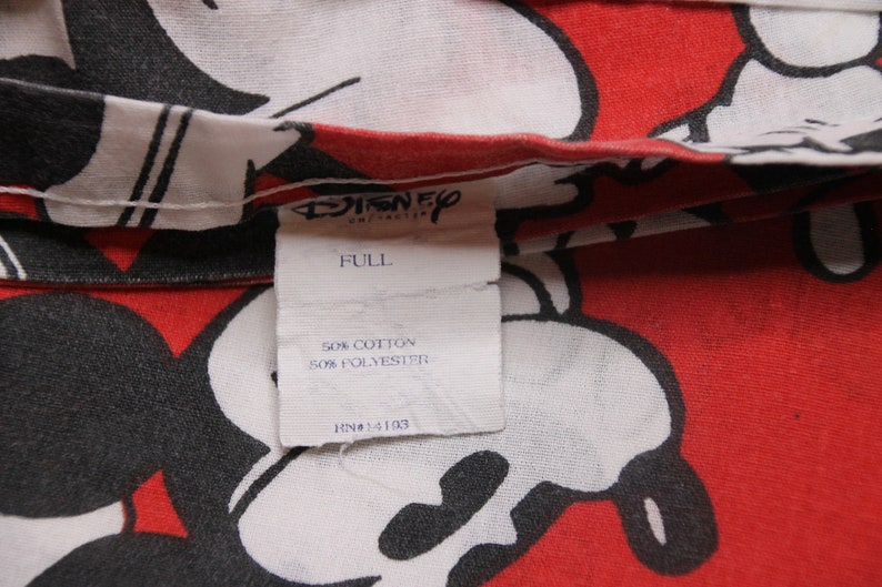 Op de afbeelding: Een wit label met zwarte tekst die "Disney", "Full", "50% Katoen 50% Polyester" en "RN#14103" luidt, tegen een rode en witte achtergrond met een Mickey Mouse-print.