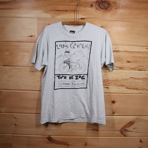 Peut inclure: Un t-shirt gris avec une illustration en noir et blanc d'une personne à vélo. L'illustration est entourée d'une bordure noire et comprend le texte "Louis Gar'eau" et "Tour de Zog". Le texte sous l'illustration se lit comme suit : "Top athletes from around the globe 10 Stages 1 Volcano Climb Overall winner to be sacrificed"