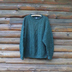 May include: A dark forest green long-sleeve jumper featuring a textured cable knit pattern throughout the front. The garment has a round crew neckline with a ribbed collar and a small interior label that reads GAP. It hangs on a wooden hanger.