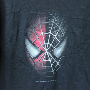May include: A black short-sleeved t-shirt featuring a graphic print of the Spider-Man mask. The mask is split vertically, with one side in red and the other in black, both overlaid with a silver web pattern. The text Spider-Man 3 is printed below.