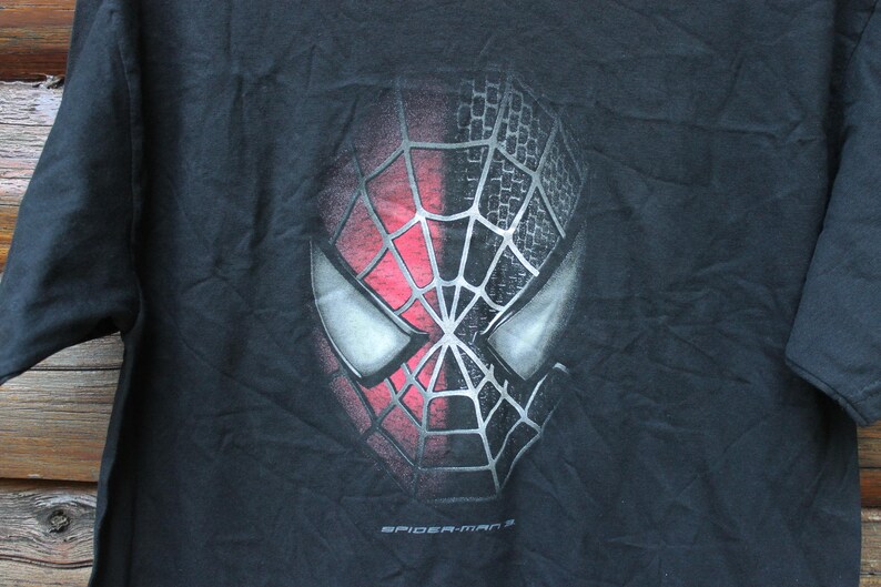 May include: A black short-sleeved t-shirt featuring a graphic print of the Spider-Man mask. The mask is split vertically, with one side in red and the other in black, both overlaid with a silver web pattern. The text Spider-Man 3 is printed below.
