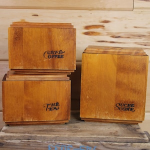 Op de afbeelding: Drie houten kannen met deksels, gelabeld met "Cafe Coffee", "The Tea" en "Sucre Sugar". De kannen staan op een houten oppervlak.