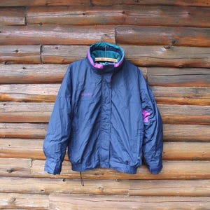 Könnte beinhalten: Eine marineblaue Columbia Jacke mit türkis- und lilafarbenem Kragen und Akzenten. Die Jacke hängt an einem Metallbügel vor einem Holzgrund. Das Columbia-Logo ist auf der Brust zu sehen.