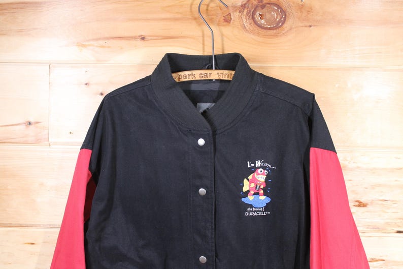 K&ouml;nnte beinhalten: Schwarze und rote Baseballjacke mit einem Cartoon-Fisch-Motiv und dem Text "I'm Waterin' for Junior Duracell".