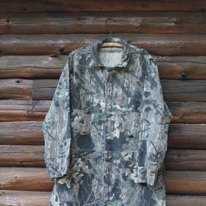 May include: A camouflage jacket hangs on a wooden hanger against a backdrop of wooden planks. The jacket features a mix of greens, browns, and greys, typical of a woodland camouflage design. It has a collared neckline, button-up front, and multiple pockets.