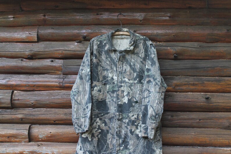 May include: A camouflage jacket hangs on a wooden hanger against a backdrop of wooden planks. The jacket features a mix of greens, browns, and greys, typical of a woodland camouflage design. It has a collared neckline, button-up front, and multiple pockets.