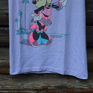 May include: A light purple t-shirt featuring a graphic of Minnie Mouse wearing a patterned top and skirt with large bows on her shoes. The design includes palm trees and the text The Walt Disney Company by Jerry Leigh, Made in U.S.A.