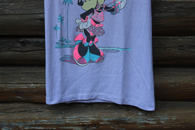 May include: A light purple t-shirt featuring a graphic of Minnie Mouse wearing a patterned top and skirt with large bows on her shoes. The design includes palm trees and the text The Walt Disney Company by Jerry Leigh, Made in U.S.A.