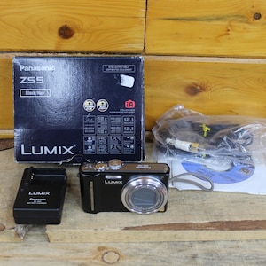 May include: A black Panasonic Lumix ZS5 digital camera with a silver lens, accompanied by its original black product box, a black battery charger, a bundled cable in a plastic bag, and a software disc, all displayed together as a complete set.