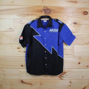 Peut inclure: Une chemise boutonnée à manches courtes noire et bleue, avec un motif d'éclair blanc. La chemise porte les logos "KERR" et "Ford". La chemise est suspendue sur un fond en bois.