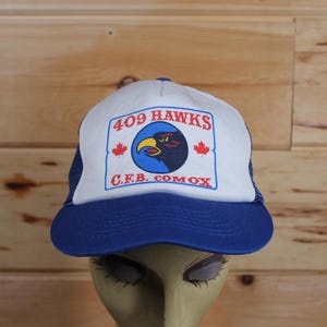 Peut inclure: Une casquette de camionneur blanche et bleue avec un écusson rouge, blanc et bleu qui dit "409 HAWKS C.F.B. COMOX" et représente un faucon noir avec un bec rouge et des yeux rouges.