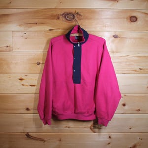 May include: A vintage pink sweatshirt with a navy blue collar and placket. The sweatshirt features a quarter-zip closure and two buttons. The garment is suspended on a metal hanger against a wooden backdrop.