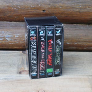 Könnte beinhalten: Ein schwarzes Box-Set mit vier WWF-VHS-Kassetten mit den Titeln SummerSlam, Over the Edge, Fully Loaded und Unforgiven. Jeder Buchrücken zeigt oben das Logo der World Wrestling Federation und unten das VHS-Etikett mit unterschiedlich farbigen Texten.