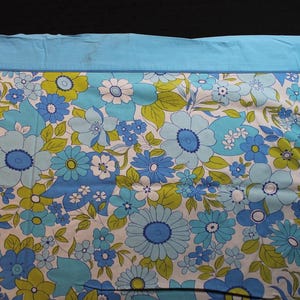 Puede incluir: Una tela floral vintage con un borde azul claro. La tela presenta un patrón de grandes flores azules, celeste y blancas con hojas verdes sobre un fondo blanco. El borde superior es de color azul claro liso.