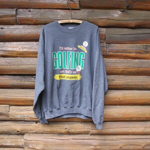 Op de afbeelding: Grijs sweatshirt met een grafische print die zegt "I'd rather be golfing and that's my final answer." De afbeelding bevat een golfbal en een golfclub.