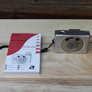 May include: A silver Canon digital camera with a black strap and a manual. The manual has a red and white cover with the text "IXUS Z50". The camera is on a light wood surface.