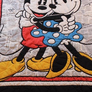 Puede incluir: Una colcha acolchada con Mickey y Minnie Mouse abraz&aacute;ndose. Los personajes est&aacute;n sobre un fondo de peri&oacute;dico. Mickey lleva pantalones cortos rojos y zapatos amarillos, mientras que Minnie luce un vestido azul de lunares. La colcha est&aacute; bordeada en rojo y negro.