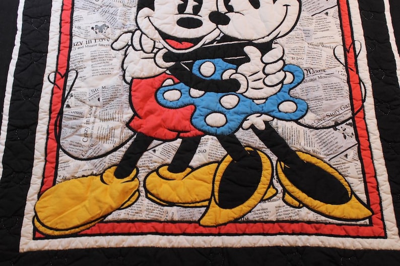 Puede incluir: Una colcha acolchada con Mickey y Minnie Mouse abraz&aacute;ndose. Los personajes est&aacute;n sobre un fondo de peri&oacute;dico. Mickey lleva pantalones cortos rojos y zapatos amarillos, mientras que Minnie luce un vestido azul de lunares. La colcha est&aacute; bordeada en rojo y negro.