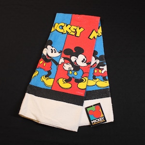 May include: Folded Mickey Mouse towel with a red, blue, and black design. The towel features Mickey Mouse in various poses, with the words "Mickey Mouse" in yellow. A tag with the Mickey Unlimited logo is attached.