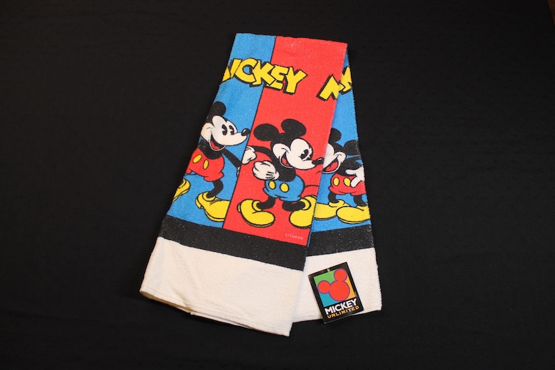 May include: Folded Mickey Mouse towel with a red, blue, and black design. The towel features Mickey Mouse in various poses, with the words "Mickey Mouse" in yellow. A tag with the Mickey Unlimited logo is attached.