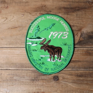 以下が含まれることがあります： 「SUCCESSFUL MOOSE HUNTER 1973」と「ONTARIO MINISTRY OF NATURAL RESOURCES」の文字が入った、緑色の楕円形のパッチ。雪景色の中にいる茶色のヘラジカが描かれています。パッチは木製の表面にあります。