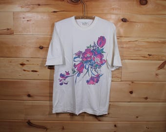 Puede incluir: Camiseta blanca con un diseño floral de tulipanes rosas y morados, detalles azules y hojas verdes. La camiseta de manga corta cuelga sobre un fondo de madera. La camiseta tiene cuello redondo.