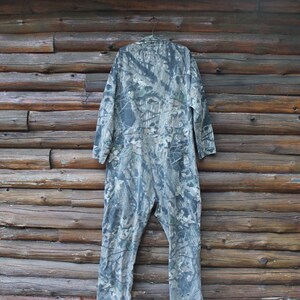 May include: A camouflage jumpsuit hangs against a wooden wall. The one-piece garment features a green, grey, and brown woodland pattern. The jumpsuit has long sleeves and a button-up front, suggesting it's designed for outdoor activities.