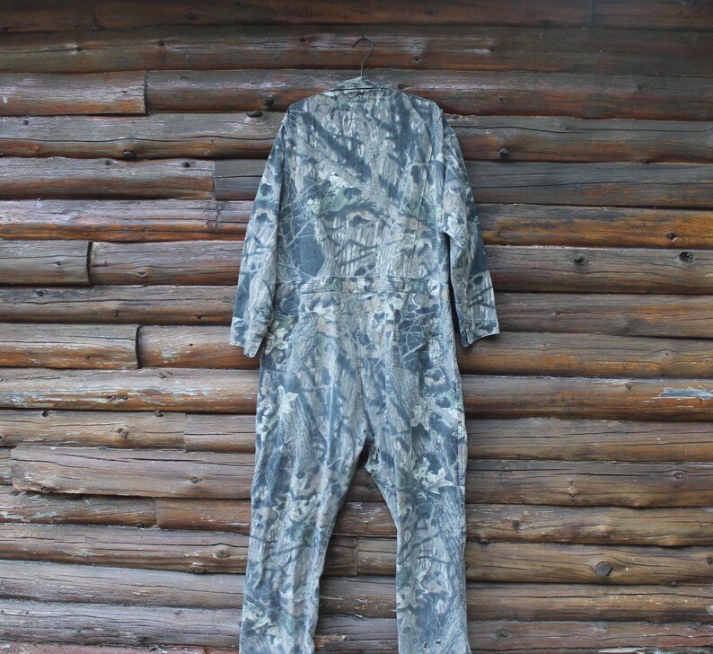 May include: A camouflage jumpsuit hangs against a wooden wall. The one-piece garment features a green, grey, and brown woodland pattern. The jumpsuit has long sleeves and a button-up front, suggesting it's designed for outdoor activities.