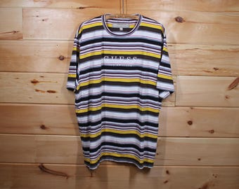 Könnte beinhalten: Ein gestreiftes T-Shirt in den Farben Weiß, Gelb, Braun und Schwarz. Das Shirt hat das Wort "GUESS" auf der Vorderseite gestickt.