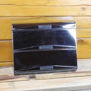 May include: A rectangular black metal bread box with a glossy finish, featuring three horizontal hinged compartments. Each compartment door has a thin, raised horizontal handle strip centered across the front surface of the unit.