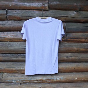 May include: A plain, light lavender short-sleeve t-shirt with a crew neckline. The garment is displayed on a wire hanger, showing the back side of the shirt. The fabric appears to be a solid, uniform color with no visible patterns, graphics, or text.