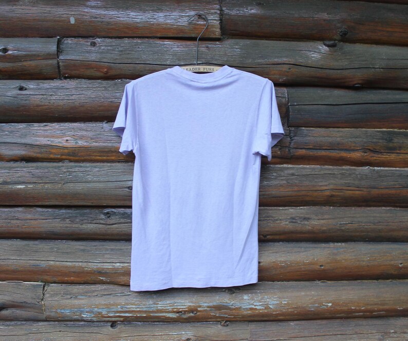 May include: A plain, light lavender short-sleeve t-shirt with a crew neckline. The garment is displayed on a wire hanger, showing the back side of the shirt. The fabric appears to be a solid, uniform color with no visible patterns, graphics, or text.