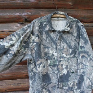 May include: A camouflage jacket with long sleeves, two front pockets, and a collared neckline. The jacket is suspended on a wooden hanger against a rustic wooden backdrop. The pattern features green, brown, and grey tones.