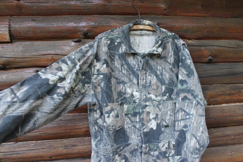 May include: A camouflage jacket with long sleeves, two front pockets, and a collared neckline. The jacket is suspended on a wooden hanger against a rustic wooden backdrop. The pattern features green, brown, and grey tones.