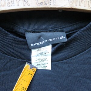 May include: A black t-shirt featuring a Spider-Man 3 branded neck label and a white care instruction tag. A yellow measuring tape is positioned against the tag, showing a measurement of approximately 3.5 inches in length for the tag area.