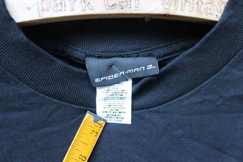 May include: A black t-shirt featuring a Spider-Man 3 branded neck label and a white care instruction tag. A yellow measuring tape is positioned against the tag, showing a measurement of approximately 3.5 inches in length for the tag area.