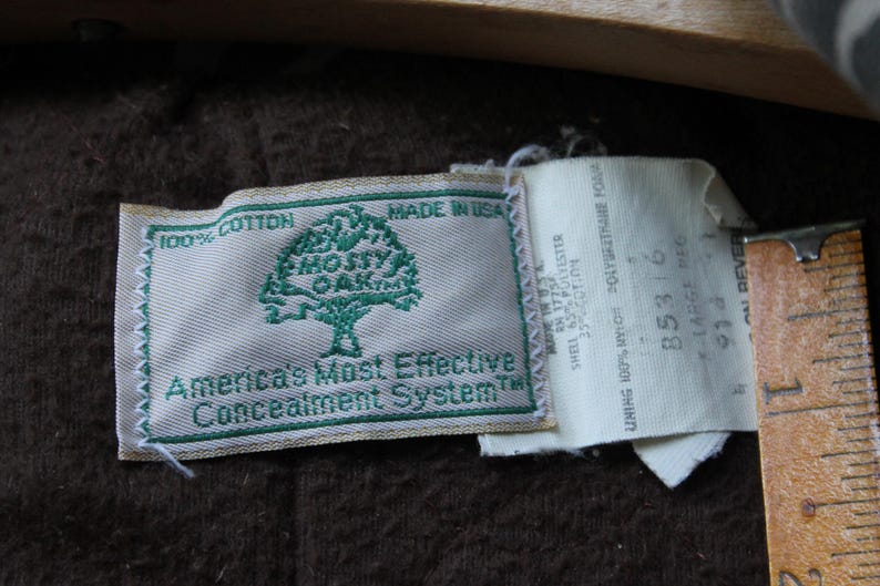 Puede incluir: Primer plano de dos etiquetas de tela. La primera etiqueta es rectangular con un borde verde y el texto "100% Algod&oacute;n" y "America's Most Effective Concealment System". La segunda etiqueta tiene texto y n&uacute;meros.